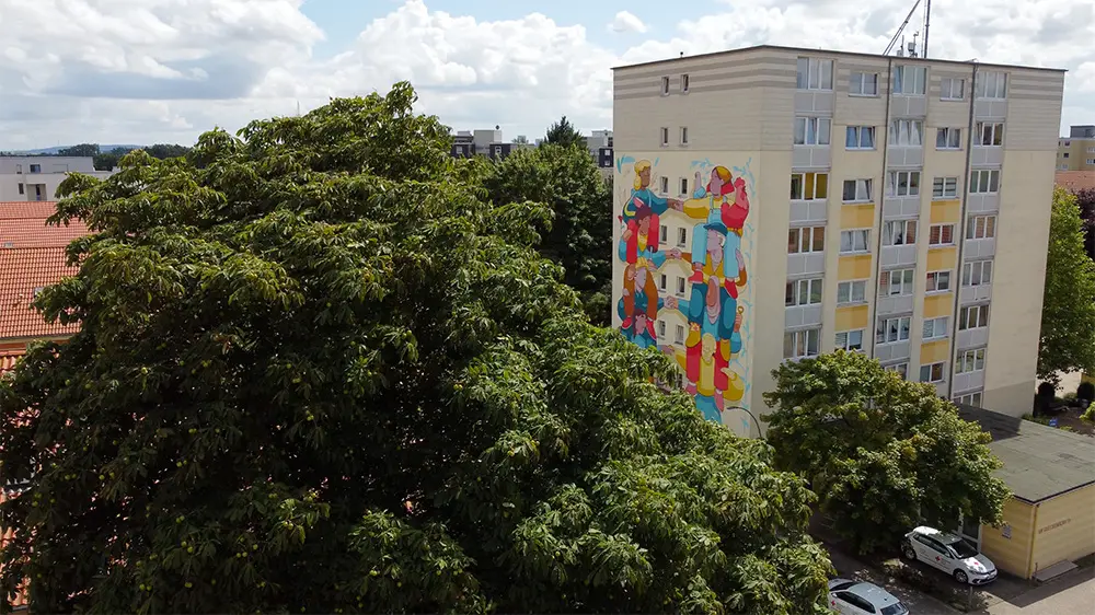 Ein Wohnhaus mit einer bunten Wandmalerei an der Seite steht zwischen grünen Bäumen und geparkten Autos unter einem teilweise bewölkten Himmel. Die Wandmalerei zeigt abstrakte Formen und leuchtende Farben.
