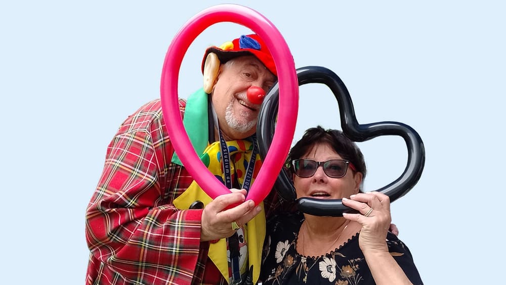 Bild: © WIEDERAUFBAU Ein Clown und eine Mieterin halten Luftballonherzen in die Kamera