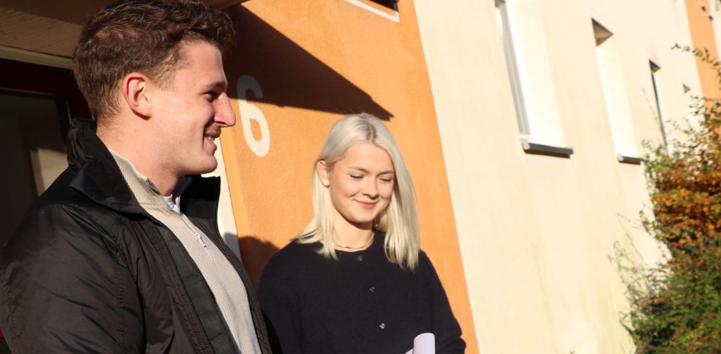 Bild: © WIEDERAUFBAU Eine Mitarbeiterin und ein Mitarbeiter im Gespräch mit einer Frau vor einem Hauseingang.