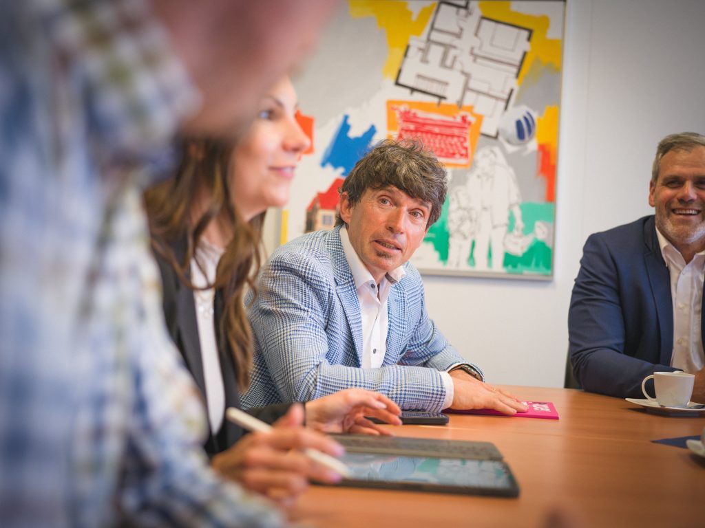 Vier Personen sitzen an einem Konferenztisch und diskutieren, während ein Mann in einer blau karierten Jacke aufmerksam zuhört. An der Wand hinter ihnen ist ein buntes abstraktes Kunstwerk zu sehen.