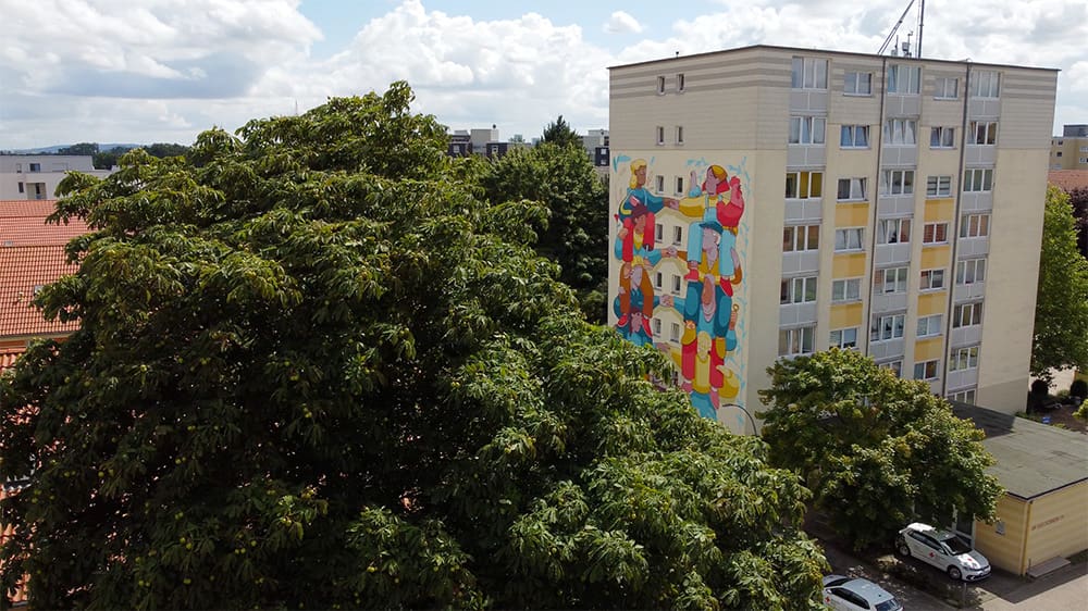 Ein Wohnhaus mit einer bunten Wandmalerei an der Seite steht zwischen grünen Bäumen und geparkten Autos unter einem teilweise bewölkten Himmel. Die Wandmalerei zeigt abstrakte Formen und leuchtende Farben.