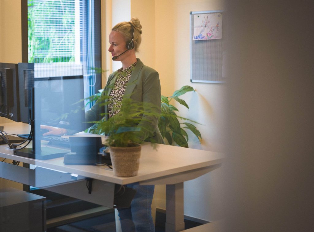 Bild: © Tom Tautz Eine Mitarbeiterin steht in ihrem Büro vor dem PC und hat ein Headset auf dem Kopf.