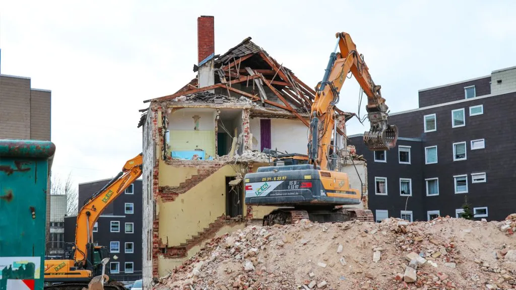 Bild: © WIEDERAUFBAU Ein Bagger steht auf einem Berg schutt und reißt ein Wohngebäude ein.