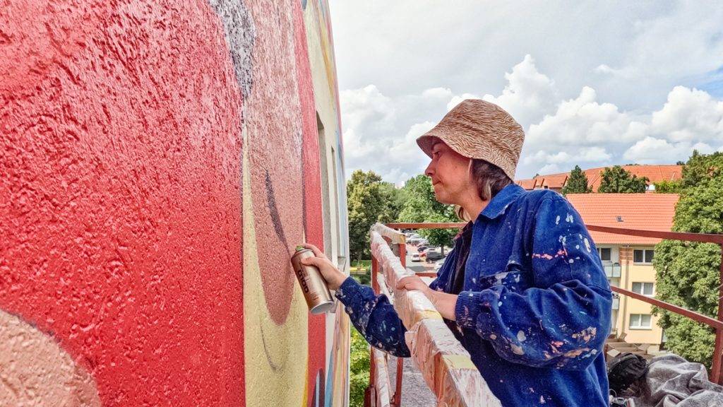 Bild: © Verena Meier Eine Frau steht auf einem Hubwagen und sprüht mit einer Spraydose ein Mural an eine Hauswand.