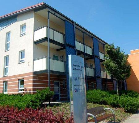 Bild: © WIEDERAUFBAU Im Hintergrund ist das dreistöckige Wohngebäude sehen, im Vordergrund steht die dazugehörige Infotafel des Betreuten Wohnen Volkmarode.