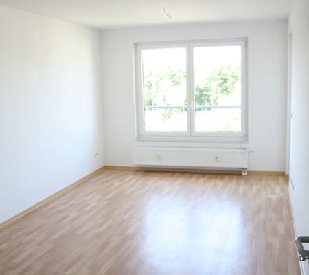 Bild: © WEDERAUFBAU Ein Wohnzimmer mit großen Fenstern, Heizung und hellbraunem Bodenbelag.