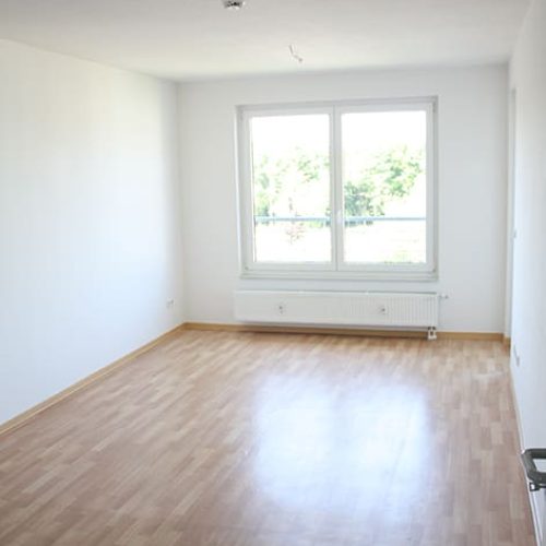 Bild: © WEDERAUFBAU Ein Wohnzimmer mit großen Fenstern, Heizung und hellbraunem Bodenbelag.