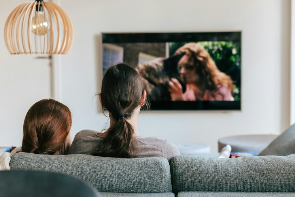 Bild: © Kevin Woblick auf Unsplash. Zwei Frauen sitzen auf der Couch und schauen auf einen Fernseher